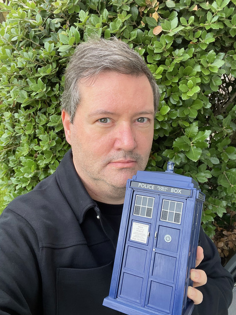 Martin, a white man with grey hair and eyes, wearing a black jacket, holding a blue Tardis prop in front of a green hedge background