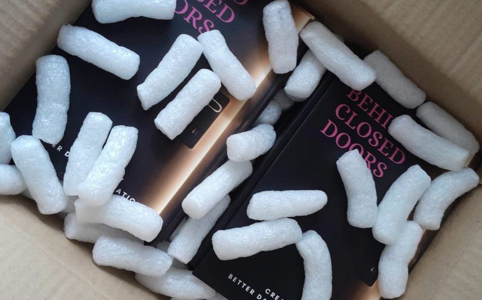 Image of Book 'Behind Closed Doors' In a cardboard box with styrofoam peanuts on top. The cover of the book is a back door slightly ajar with pink text.