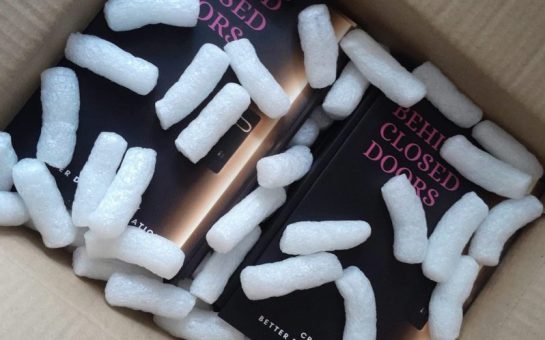 Image of Book 'Behind Closed Doors' In a cardboard box with styrofoam peanuts on top. The cover of the book is a back door slightly ajar with pink text.