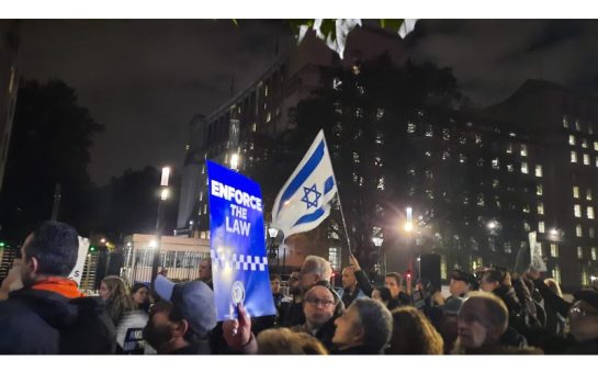 A protestor waving an Israeli flag in a crowd with someone else holding up a sign that reads 'Enforce the Law'