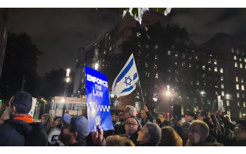 A protestor waving an Israeli flag in a crowd with someone else holding up a sign that reads 'Enforce the Law'