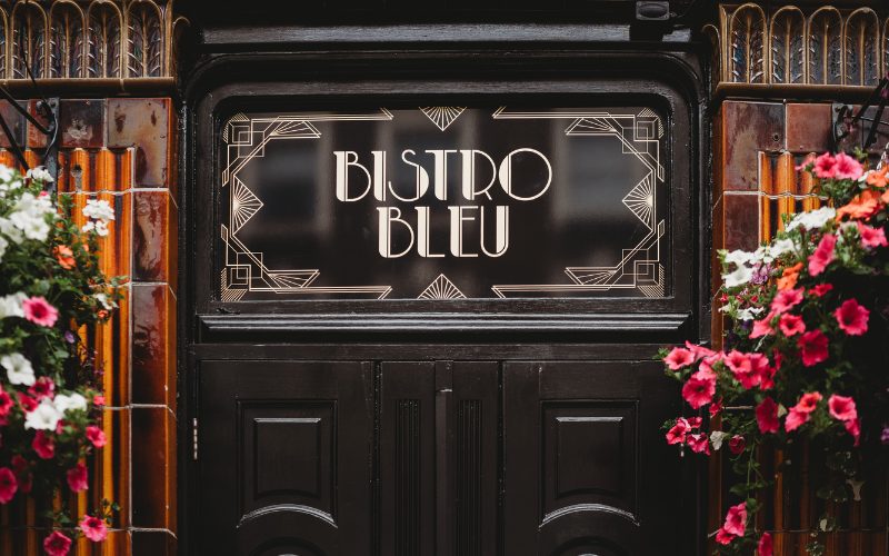 A picture of the entrance to Bistro Bleu, with an art deco logo and hanging baskets either side of it with pink and yellow flowers
