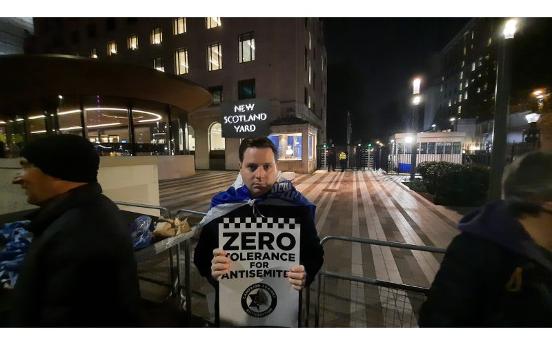 A man holding a sign that reads 'Zero tolerance for antisemites', wearing an Israeli flag, standing in front of the New Scotland Yard revolving sign