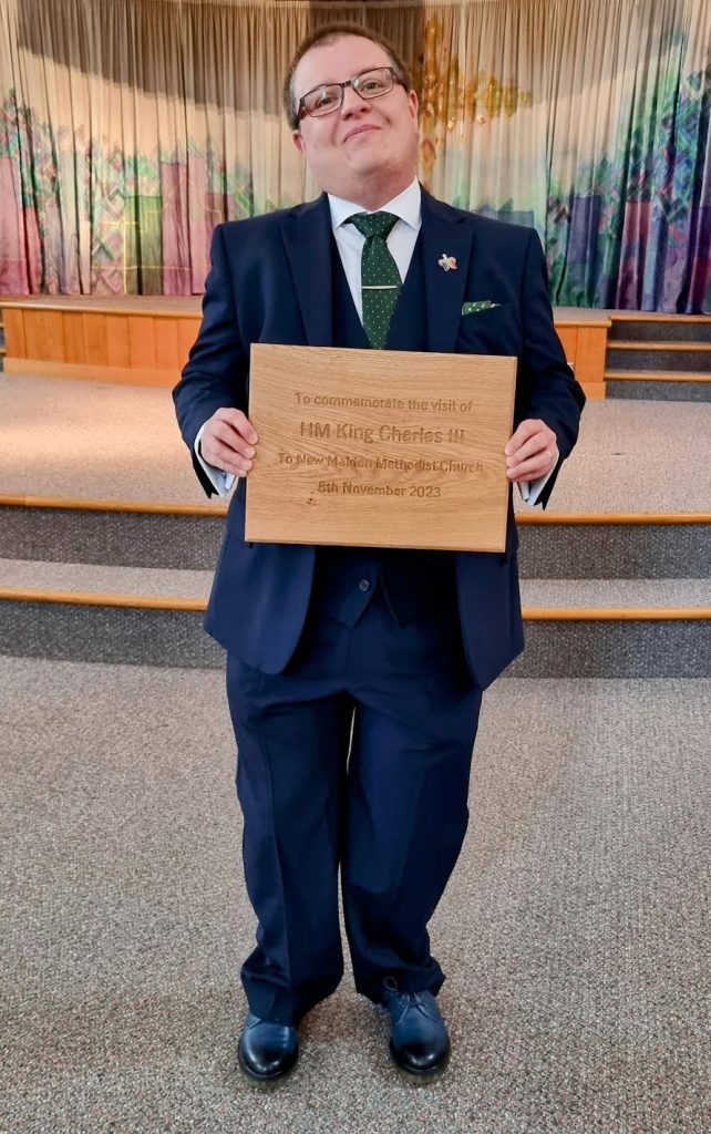 Church Minister stands with wooden plaque inscribed with 'To commemorate the visit of HM King Charles III to New Malden Methodist Church 8th November 2023'