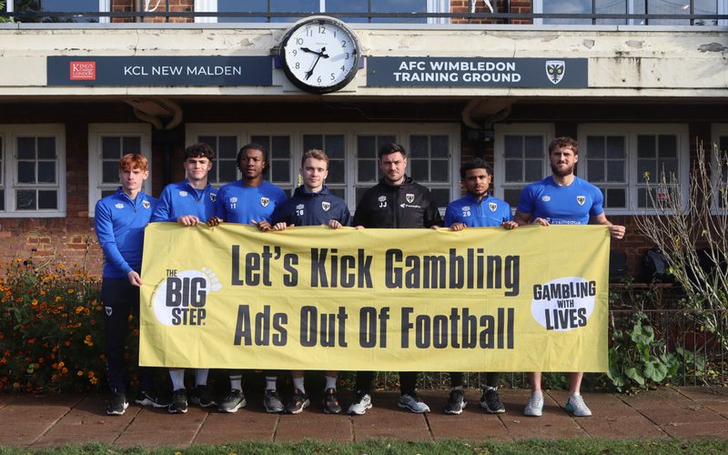 AFC Wimbledon holding up a sign produced by the Big Step campaign which reads: "Let's Kick Gambling Ads Out Of Football"