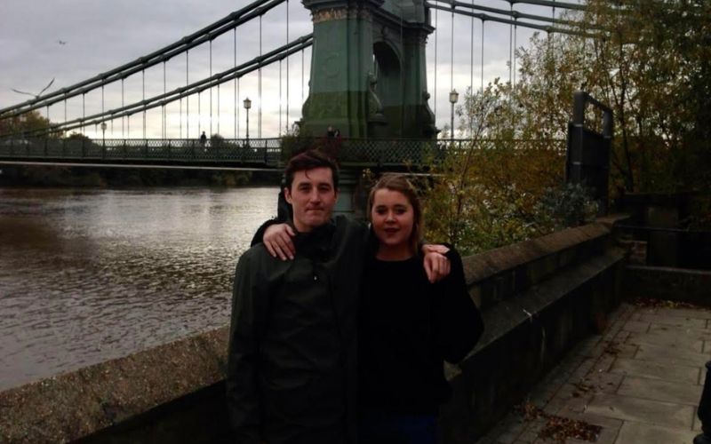 A picture of Tom Harding and his sister Katy with their arms round each other next to Hammersmith Bridge on a rainy day