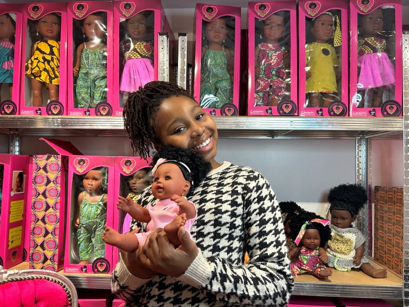 A young girl holds a black doll in front of a toy store shelf filled with black dolls