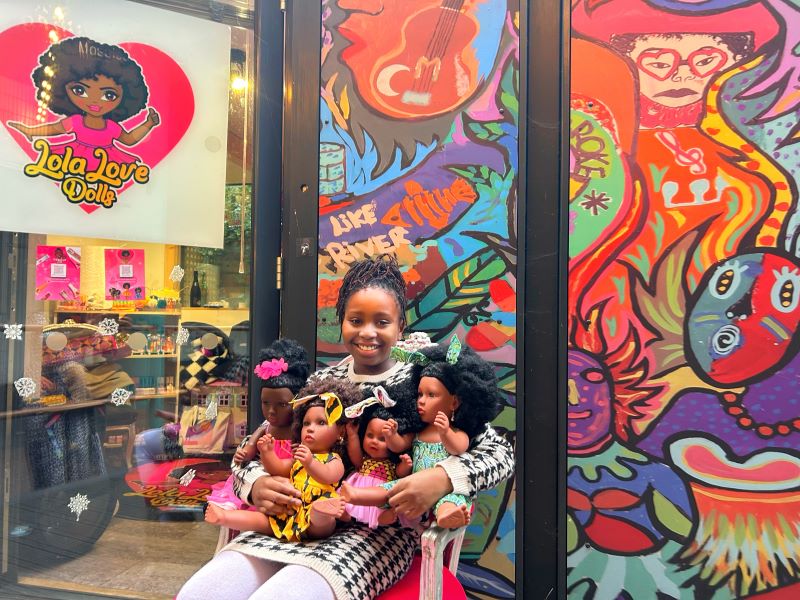 A young girl in front of a toy store (Lola Love Dolls) holding for black dolls 