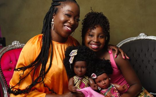 A mother and daughter sit with two black dolls between them. The dolls have varying afros and skin tones.