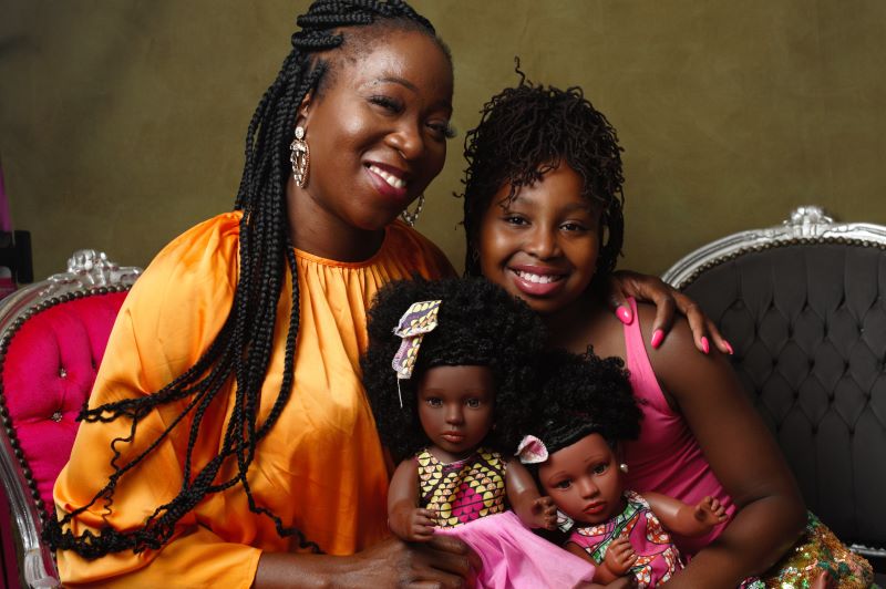 A mother and daughter sit with two black dolls between them. The dolls have varying afros and skin tones.