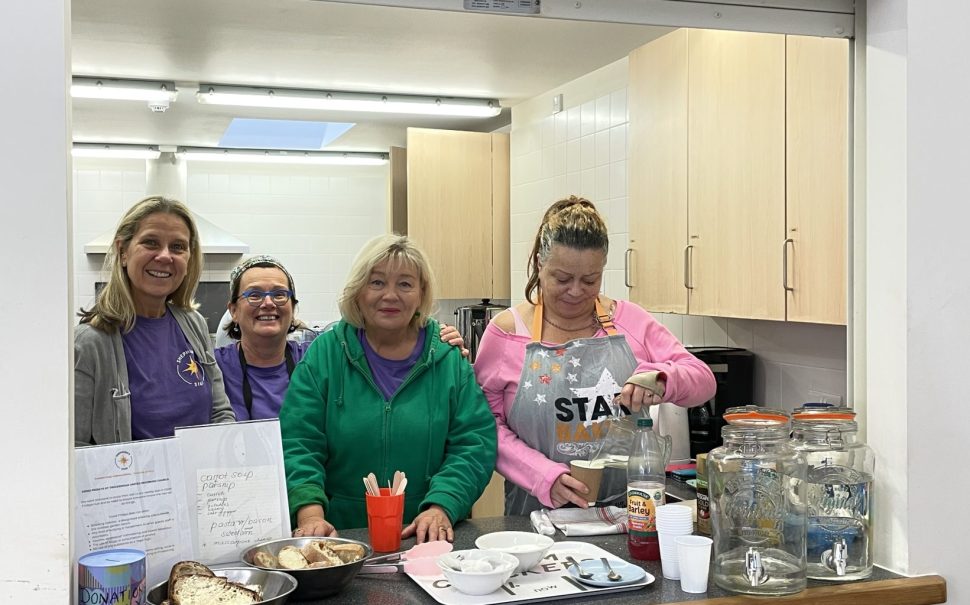 Image shows four women stood together smiling in a community kitchen set up by charity Shepherd's Star. The fourth woman on the right is pouring a drink.