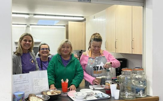 Image shows four women stood together smiling in a community kitchen set up by charity Shepherd's Star. The fourth woman on the right is pouring a drink.