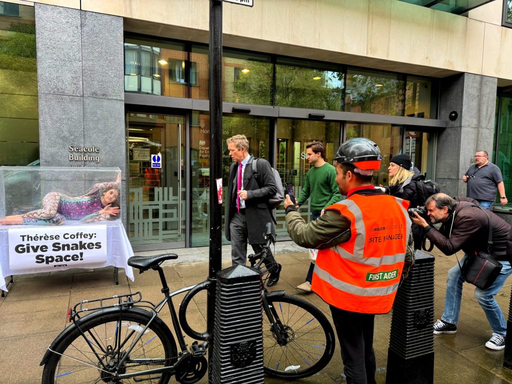 Woman dressed in colourful snake suit in a small rectangular glass tank outside the DEFRA building, with bystanders photographing her on their cell phones and cameras.