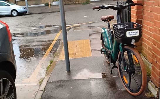 An e-bike which is discarded on the pavement. The route to pass is very narrow because there is a signpost on the other side of the pavement.