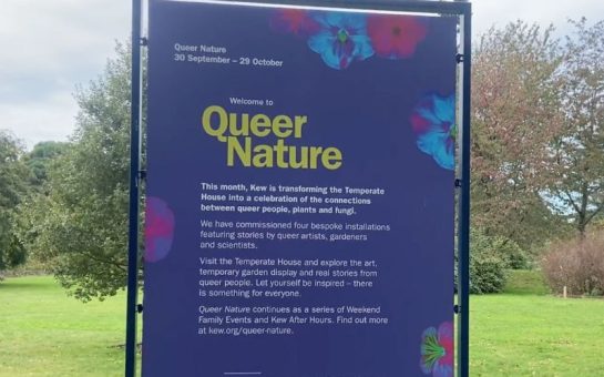 The opening sign for the Queer Nature exhibit. It is blue with yellow text which says welcome to Queer Nature, and the border is decorated with flowers. The rest of the bolded text says "this month, Kew Gardens is transforming the Temperate House into a celebration of the connections between queer people, plants and fungi.