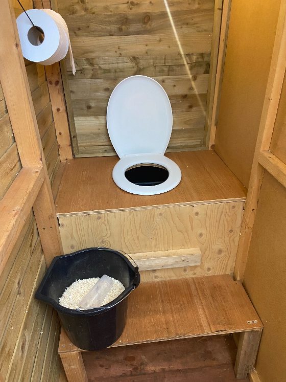 A compost toilet in a wooden hut with a bucket of sawdust next to it.