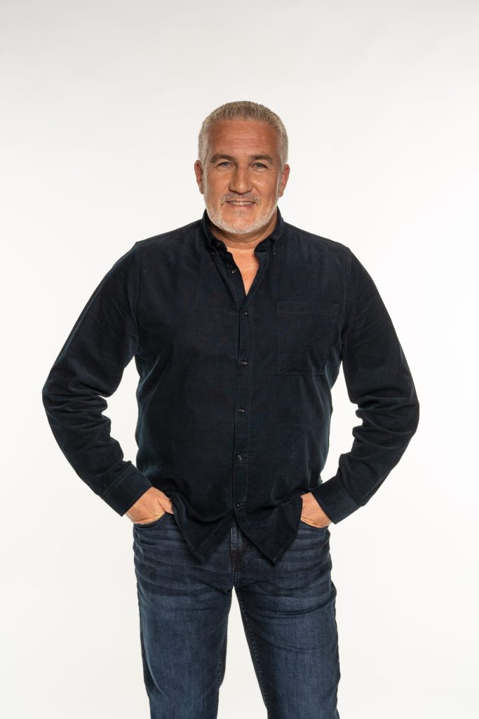 GBBO's Paul Hollywood stands in a shirt and jeans with his hand in his pockets smiling to the camera. 