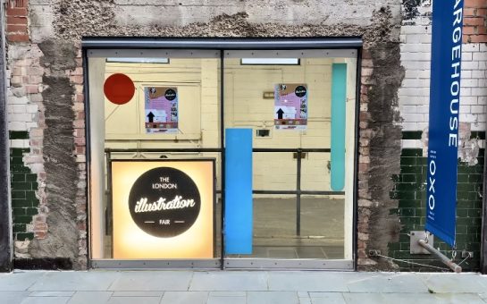 Glass window looking into the London Illustration Fair. The London Illustration Fair logo is displayed on a light box in the window.