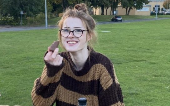 A photo released by Cheshire Constabulary of Brianna Ghey, she is sat at a park bench smiling towards the camera and wearing a brown stripey knit jumper, her hair is in a messy bun and she is wearing a pair of black glasses