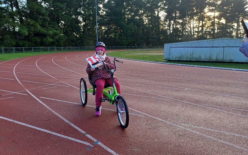 Jessica, a woman, on a green recumbent tricycle holding a sign saying "15 laps"