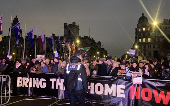 A picture of the crowd at Parliament Square standing behind a large poster that says "Bring Them Home Now" Standing in front of the crowd is a man from the Community Security Trust