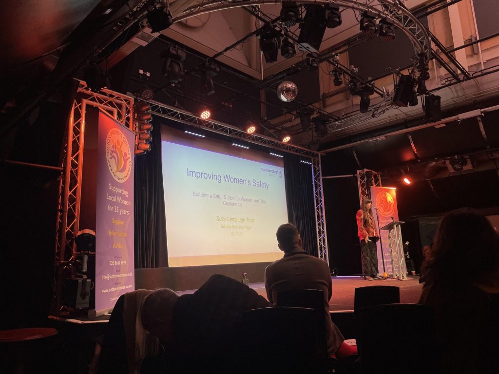 Image of the 'Building a Safer Sutton for Women and Girls' conference. A woman is talking, and the slide behind her reads "Improving Women's Safety"