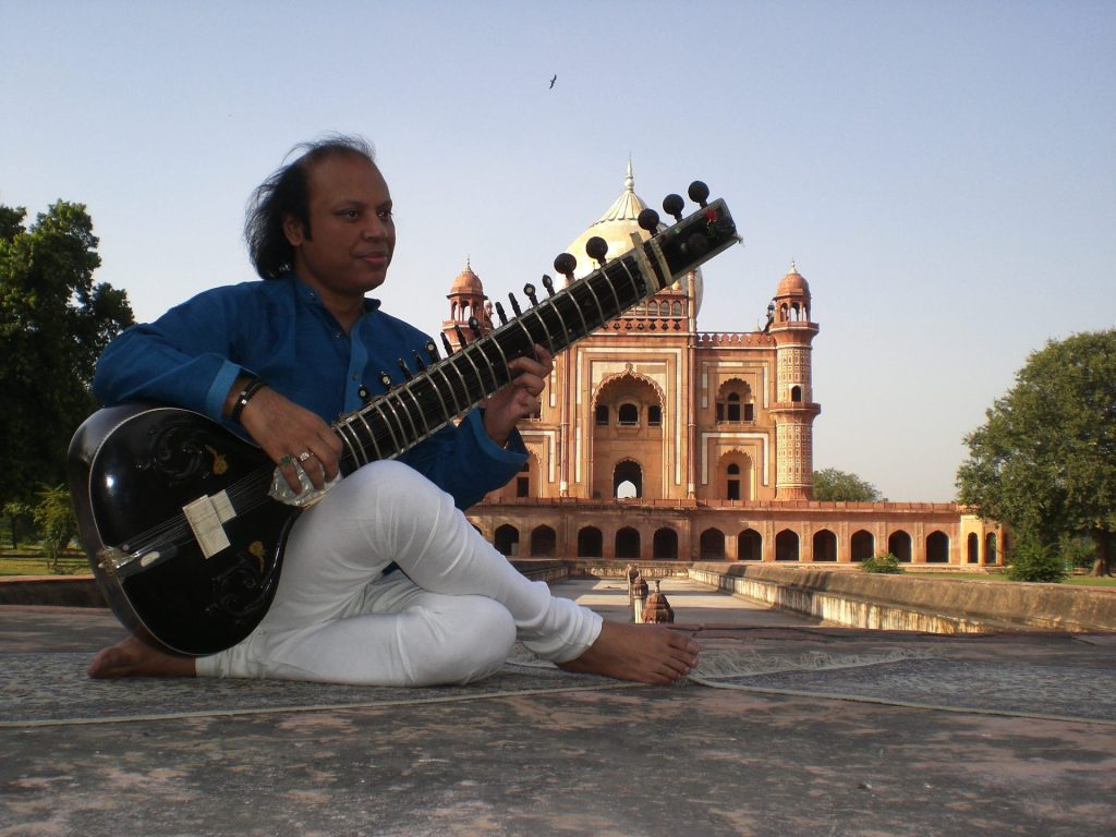 Nishat Khan sitting on the ground in front of a temple playing a sitar.