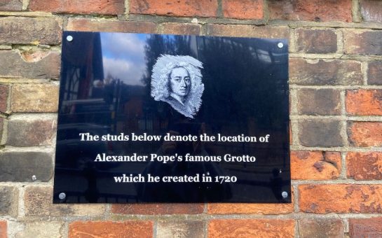 Plaque outside of Alexander Pope's Grotto. On a brick wall. Is black with white writing saying "The studs below denote the location of Alexander Pope's famous Grotto which he created in 1720." And above the writing is an etching of Alexander Pope.