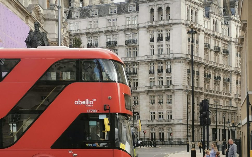 A red London bus on the left of the image, driving toward the right. In front of a old London building. Abellio, written on the side of the bus, is staging bus strikes in south-west London in January 2024.