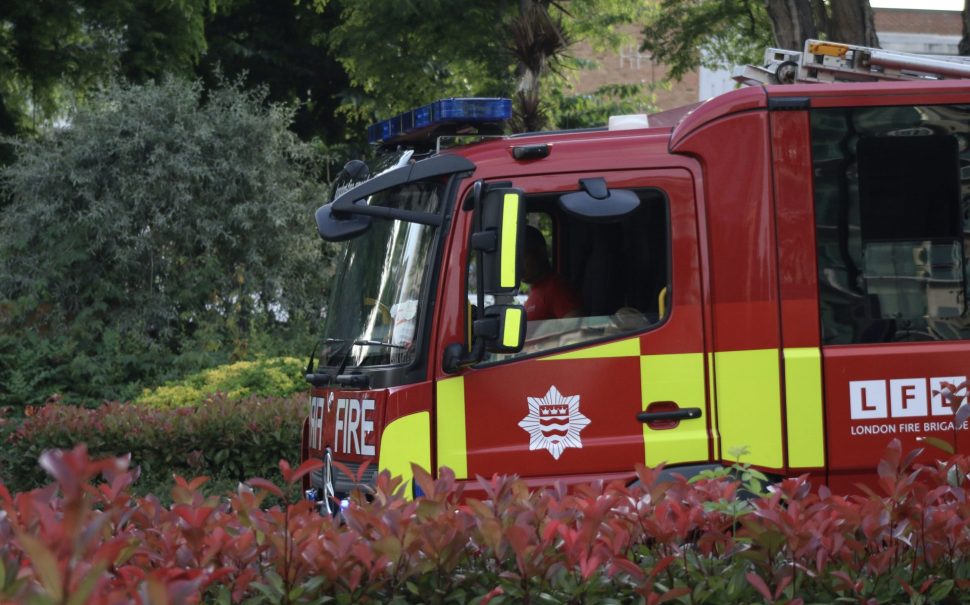A red London Fire Brigade fire engine driving from right to left, with one person in the front.