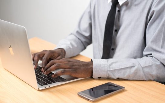 A picture of a man in a shirt an tie typing on a Macbook laptop with his phone next to him on the table