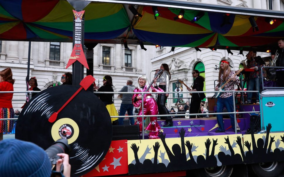 Parade Float driving past with woman in pink jumpsuit playing guitar and man with long hair also playing guitar