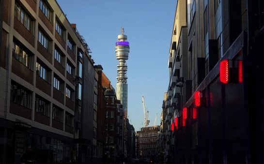 A street view of The BT Tower in blue skies, looking up at it between a row of buildings