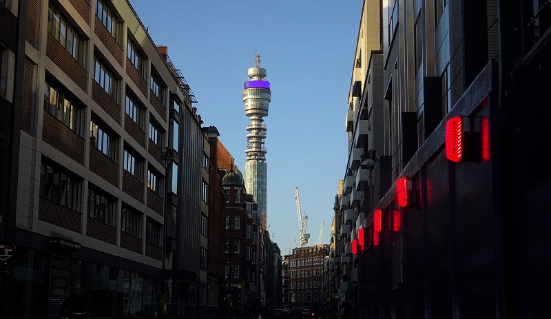 A street view of The BT Tower in blue skies, looking up at it between a row of buildings
