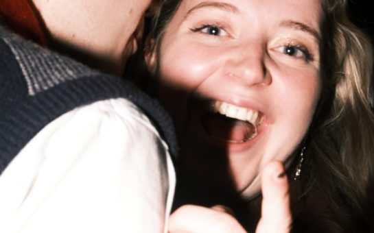 Woman looking joyful at camera smiling whilst man snuggles into her neck.