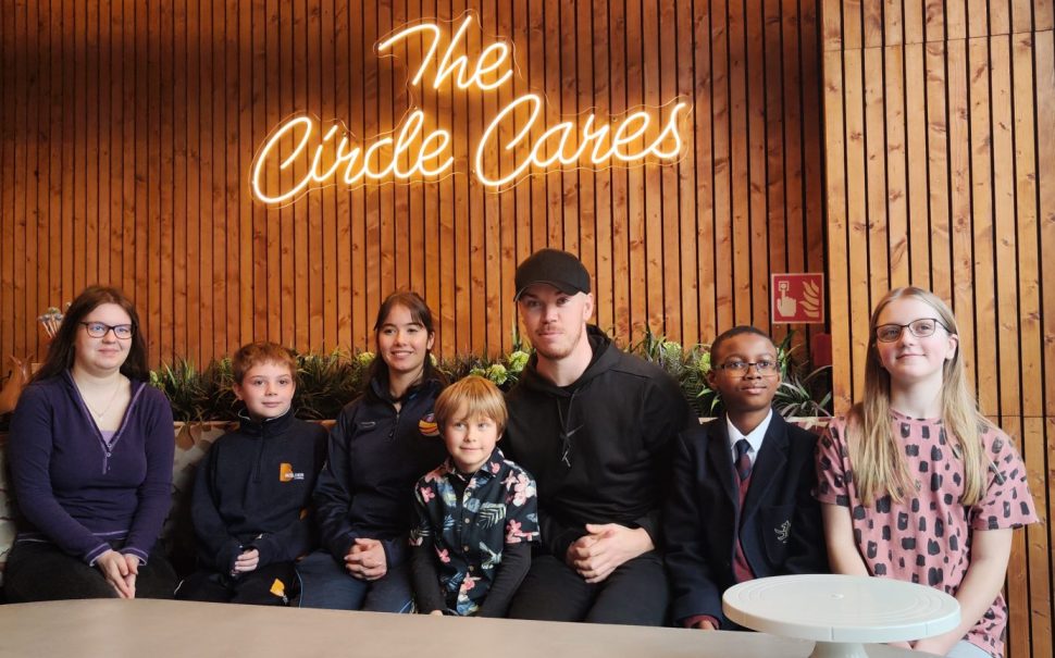 Actor Will Poulter in Circle Cafe sat at table with young children celebrating anniversary wooden wall behind with light up words The Circle Cafe