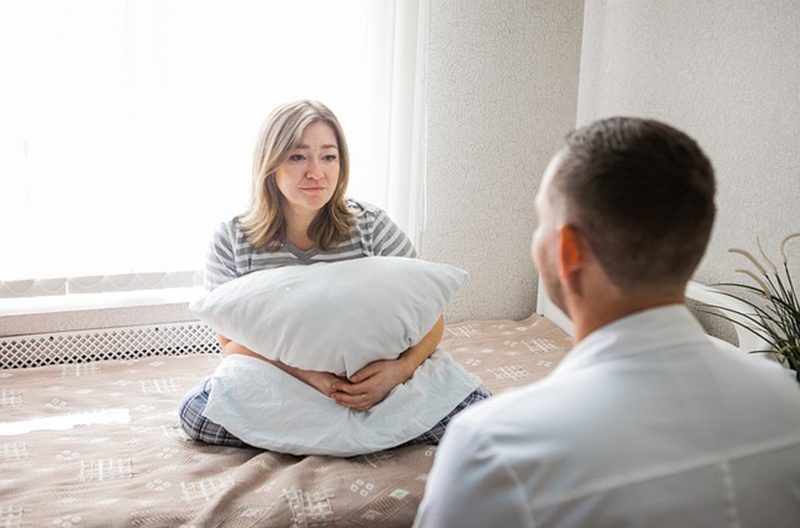 A woman, possibly suffering from mental health disorders, sits on a bed to receive some councilling