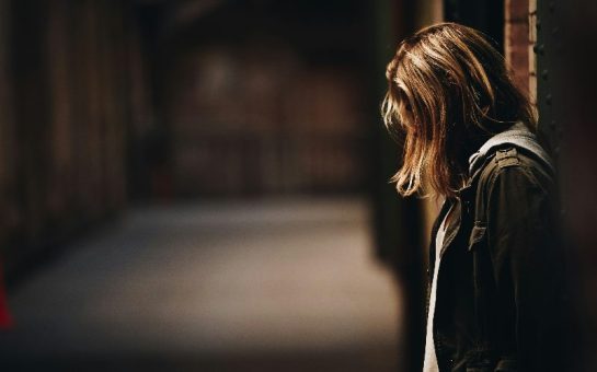 Young woman with her face covered by her hair looks down at the floor with a dark alleyway in the background