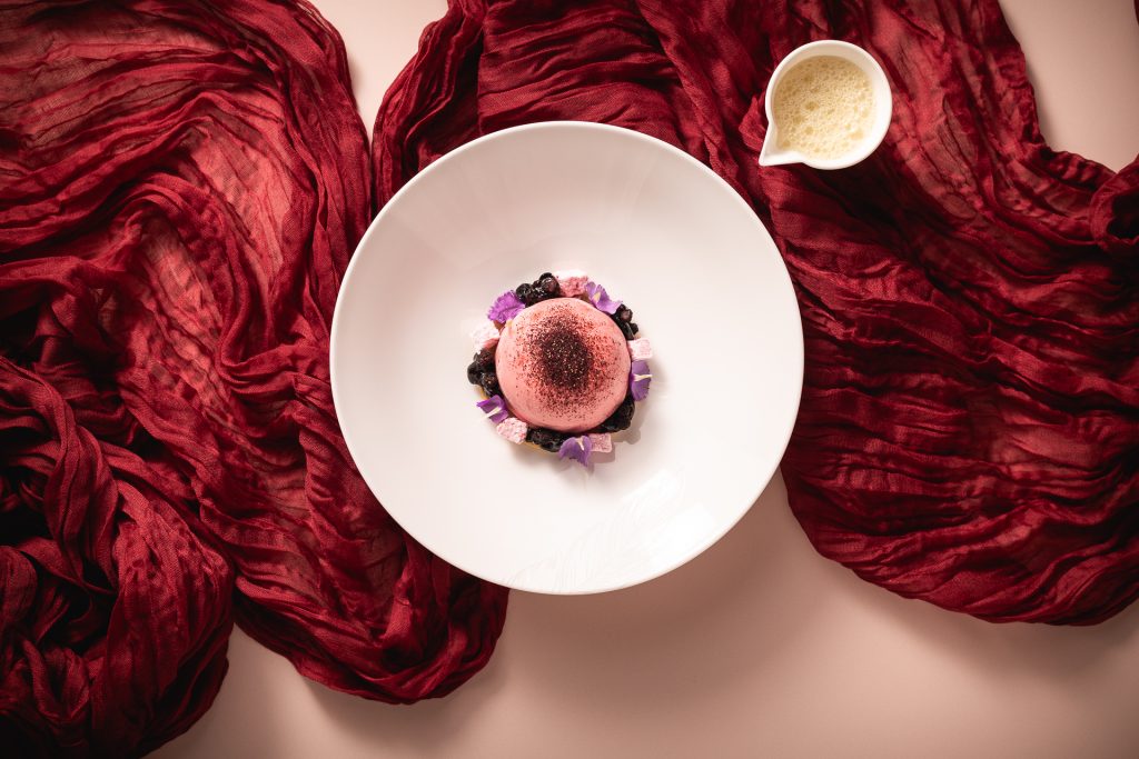 Dramatic shot of dish presented for Sally Abé's celebration of International Women's Day at The Pem. There is the main dish which includes pink and purple flowers and a sauce on the side.