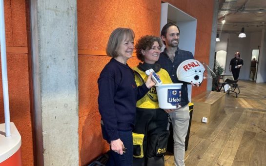 A photo of two representatives from the RNLI (Gianna Saccomani & Miranda Jaggers) with Euphonix's musical director (Thomas Chapman) holding an RNLI donations box and helmet.