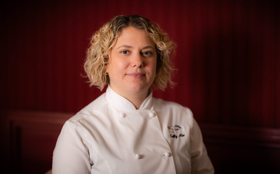 A photo of celebrated chef, Sally Abé in chef's whites. Her jacket says The Pem and her name. She is smiling. The backdrop is red.