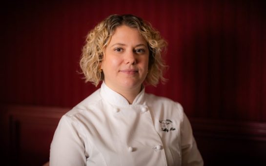 A photo of celebrated chef, Sally Abé in chef's whites. Her jacket says The Pem and her name. She is smiling. The backdrop is red.