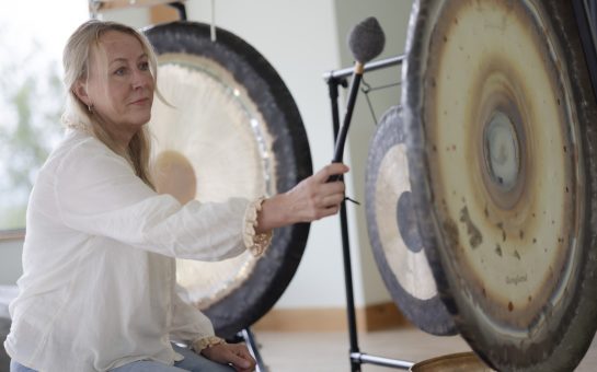 Sophie Pumfrey, gong bath practitioner, is banding a gong with a mallett in a sound bath.