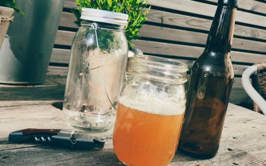 A jar of beer with a bottle on a brown wooden table outside in the sun.