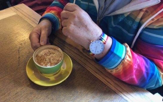 Shows a man wearing a rainbow quarter-zip jumper with a rainbow watch, drinking a coffee.