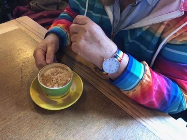Shows a man wearing a rainbow quarter-zip jumper with a rainbow watch, drinking a coffee.
