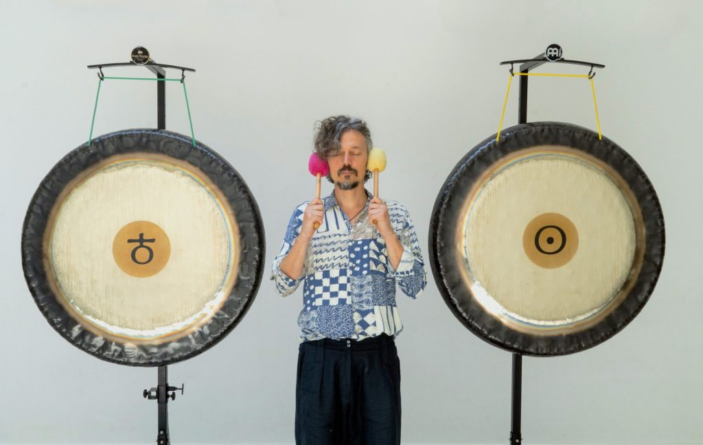 Simone Salvatici, a gong sound bath practioner, standing between two large gongs. He is holding two mallets next to his ears.