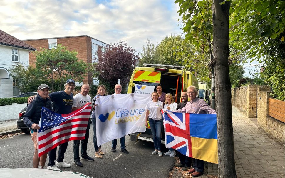 Charity Medical Life Lines Ukraine stand with flags in front of a refurbished ambulance ready to send the Ukraine. Pictured with an American Flag a flag with their charity logo and a flag that is half the UK flag and half the Ukrainian flag.