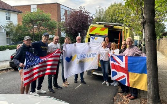 Charity Medical Life Lines Ukraine stand with flags in front of a refurbished ambulance ready to send the Ukraine. Pictured with an American Flag a flag with their charity logo and a flag that is half the UK flag and half the Ukrainian flag.