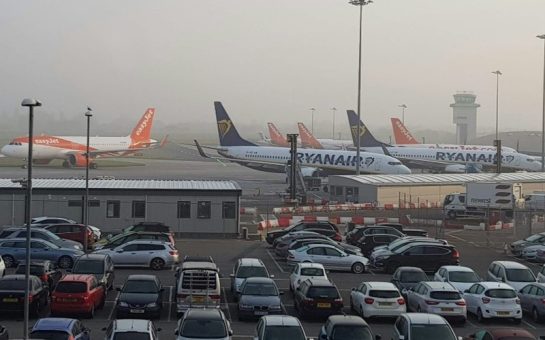 Photo of an airport carpark. Three stationary planes are in the background against a foggy sky with rows of parked cars in the foreground.